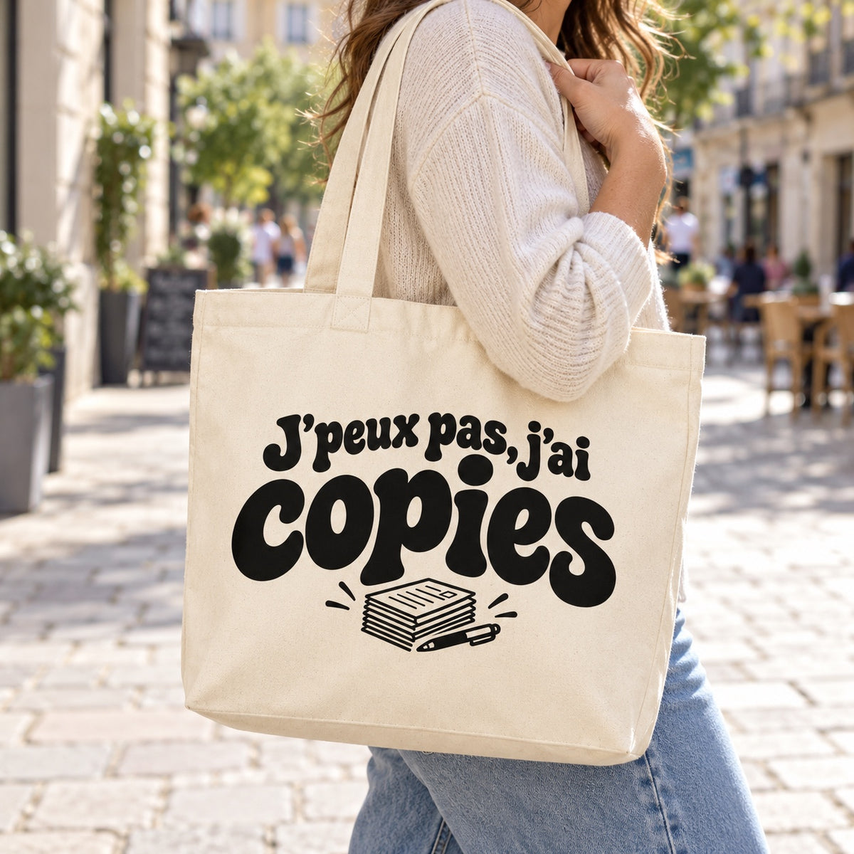 Tote bag « J'peux pas, j'ai copies » Premium porté à l'épaule dans une rue piétonne ensoleillée