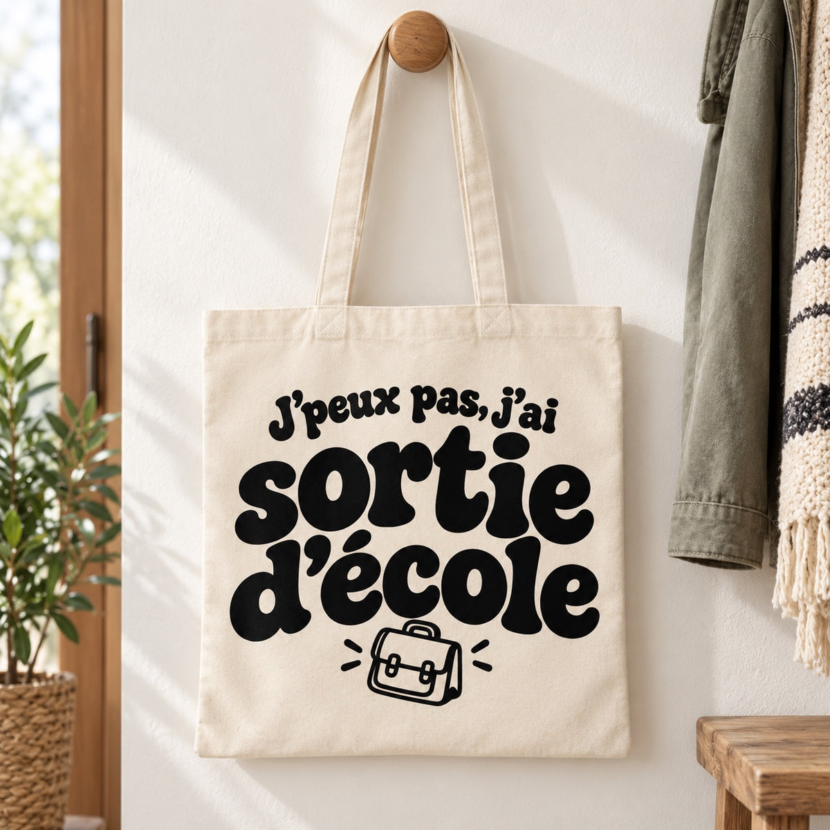 Tote bag « J'peux pas, j'ai sortie d'école » Essentiel suspendu à une patère dans une entrée lumineuse