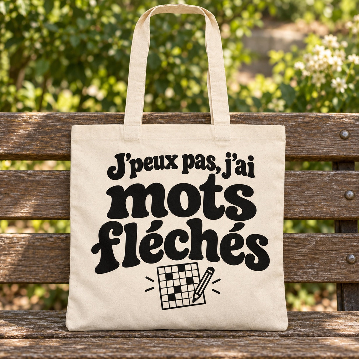 Tote bag « J'peux pas, j'ai mots fléchés » Essentiel posé sur un banc en bois dans un jardin