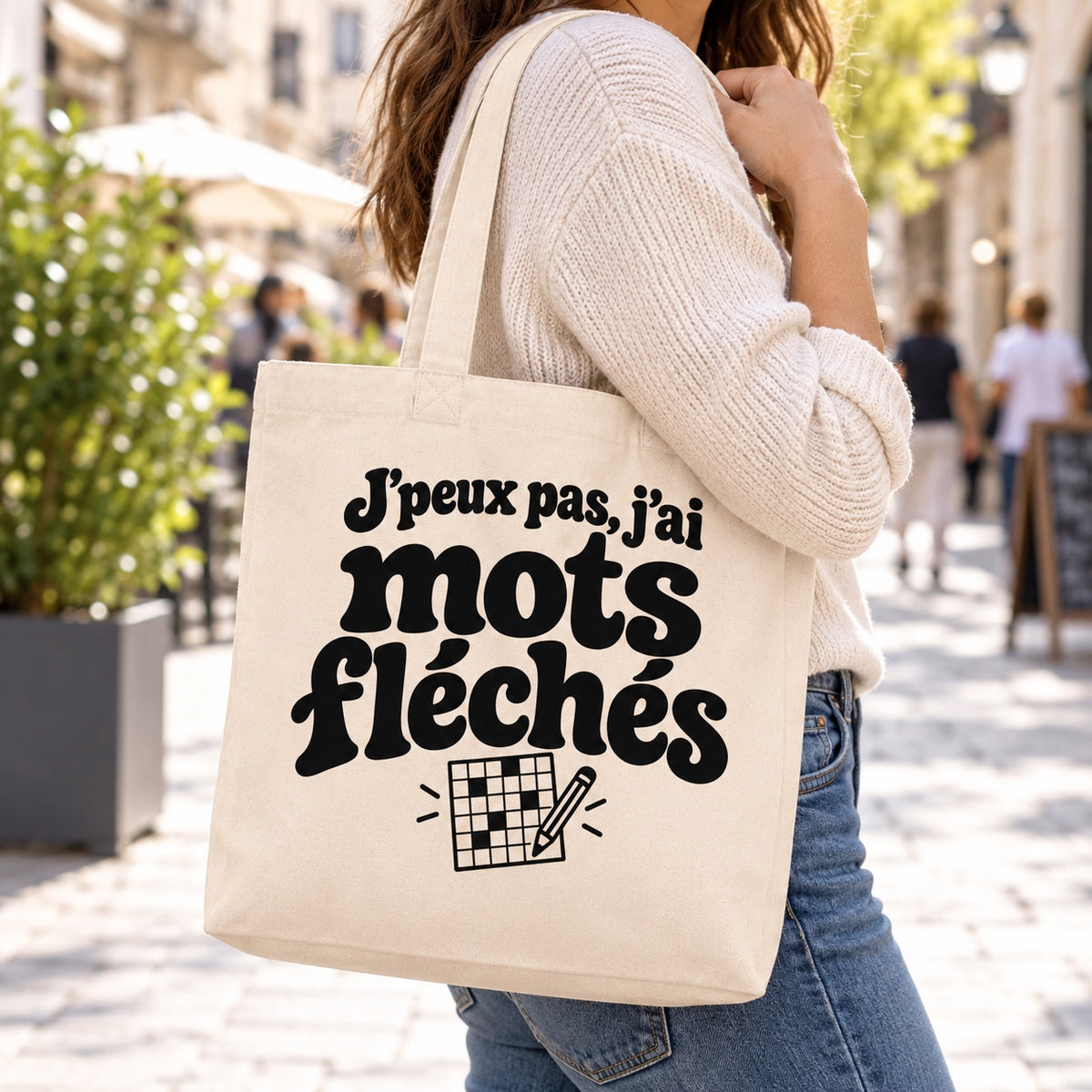 Tote bag « J'peux pas, j'ai mots fléchés » Premium porté à l'épaule dans une rue piétonne ensoleillée