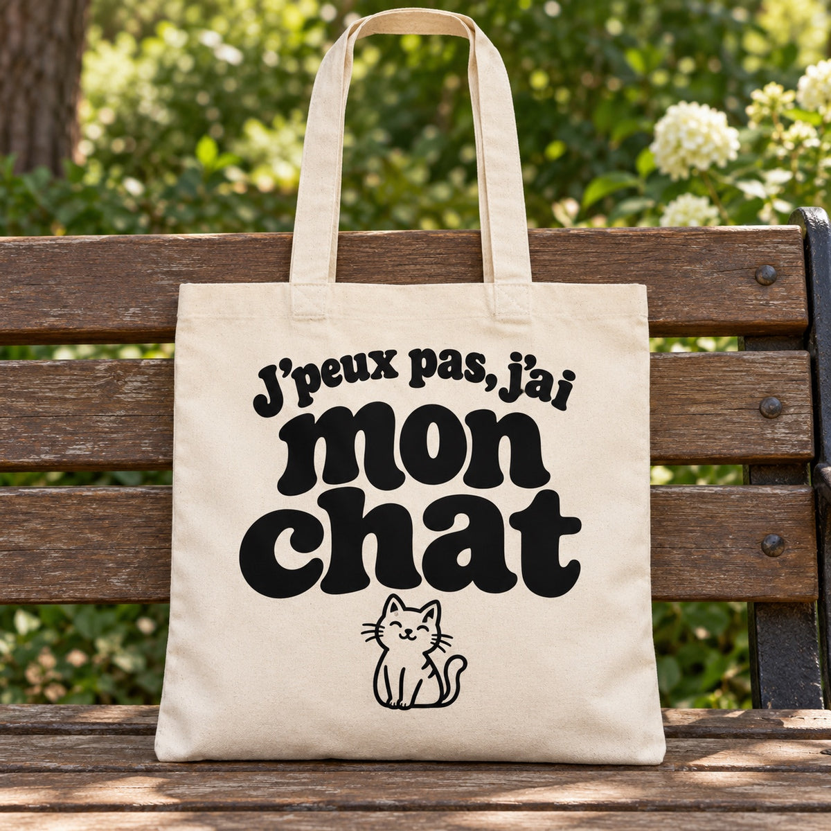 Tote bag « J'peux pas, j'ai mon chat » Essentiel posé sur un banc en bois dans un jardin