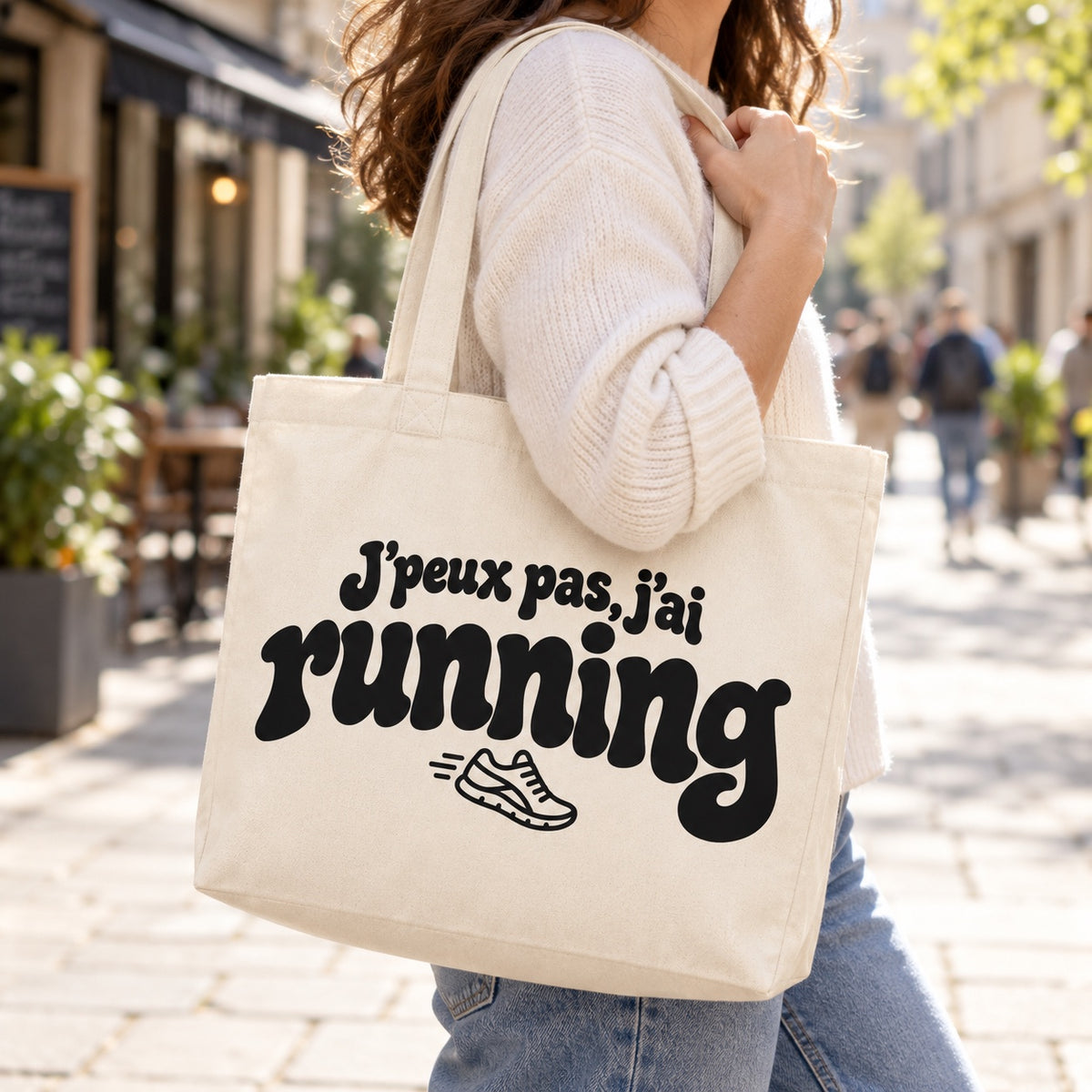 Tote bag « J'peux pas, j'ai running » Premium porté à l'épaule dans une rue piétonne ensoleillée