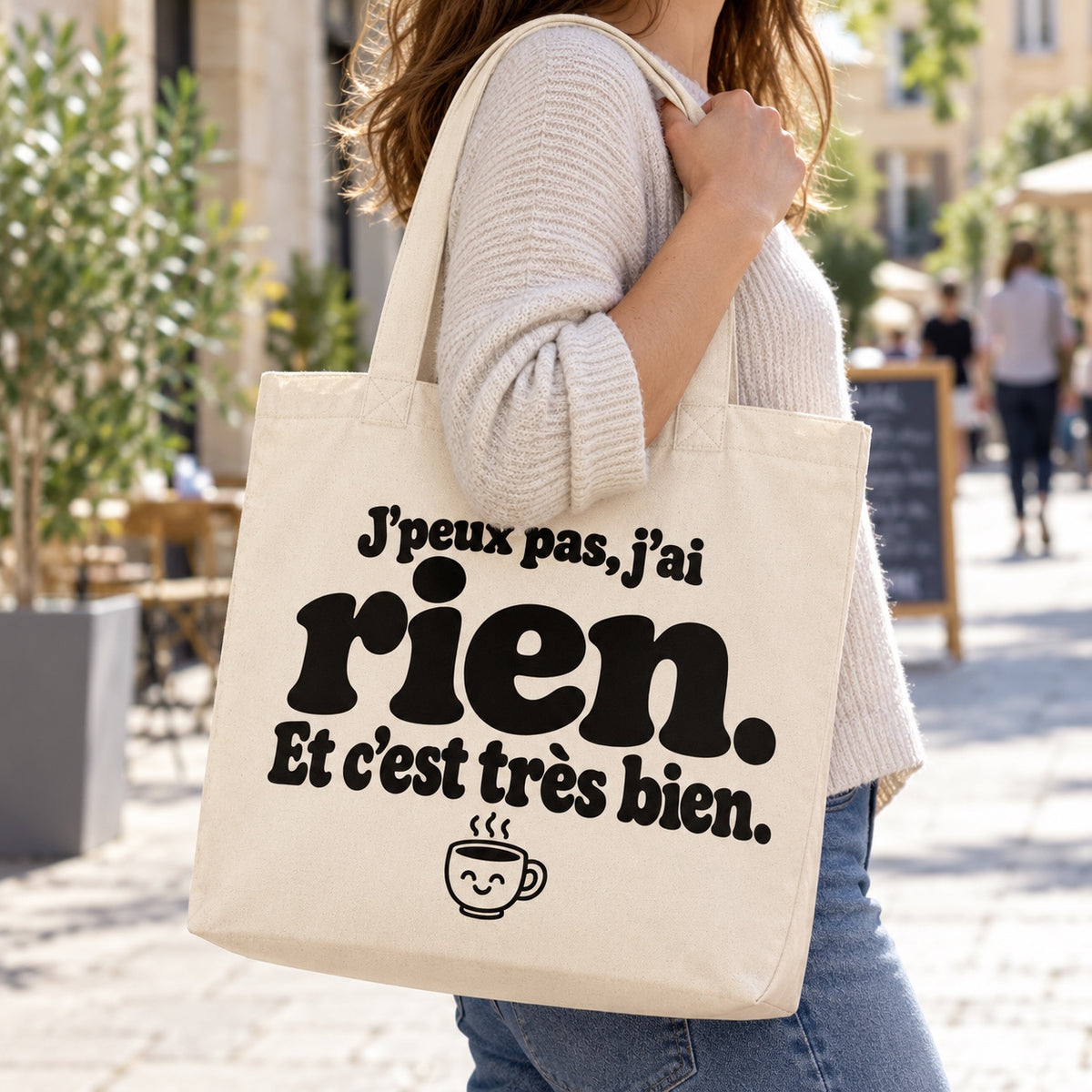 Tote bag « J'peux pas, j'ai rien. Et c'est très bien. » Premium porté à l'épaule dans une rue piétonne ensoleillée