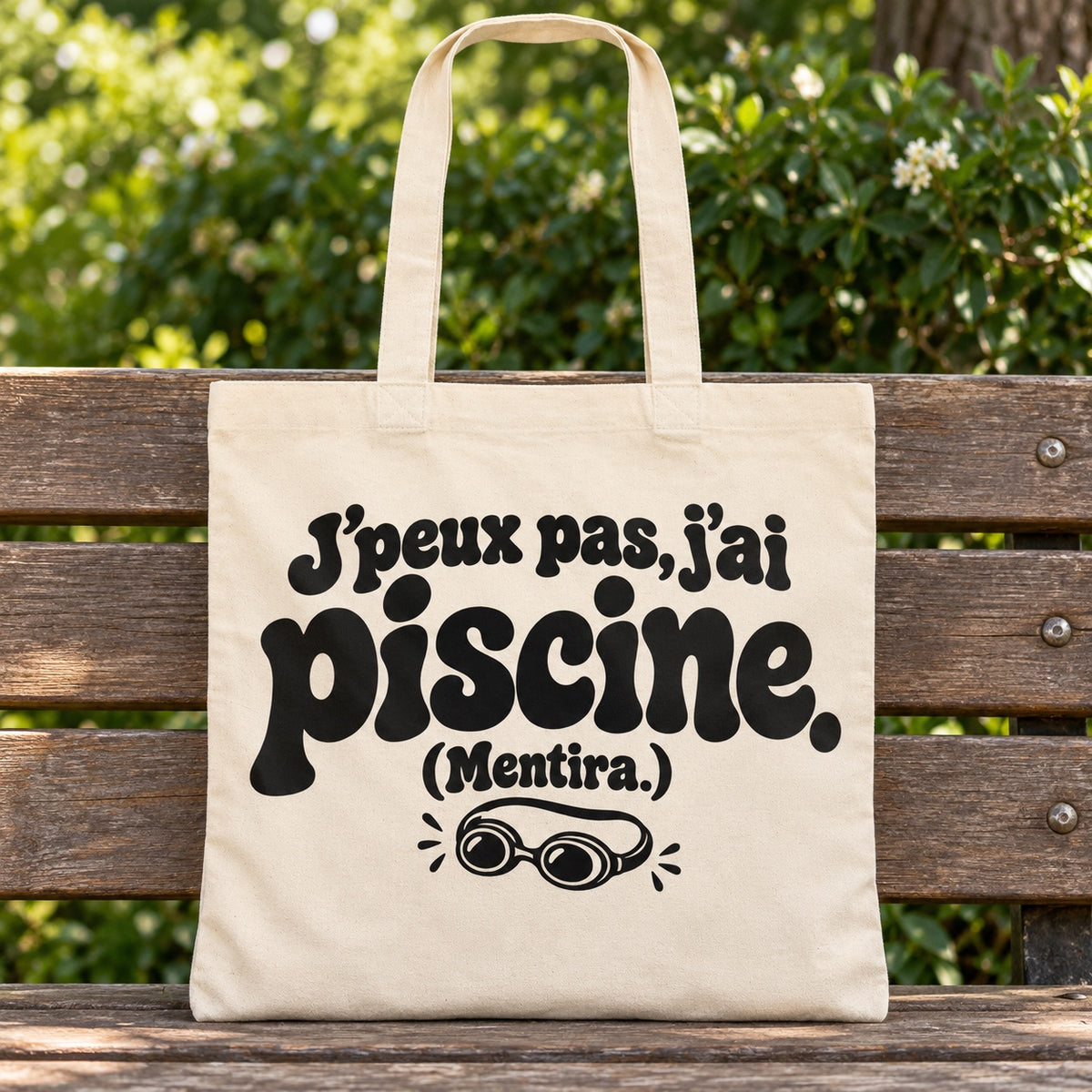 Tote bag « J'peux pas, j'ai piscine. (Mentira.) » Essentiel posé sur un banc en bois dans un jardin