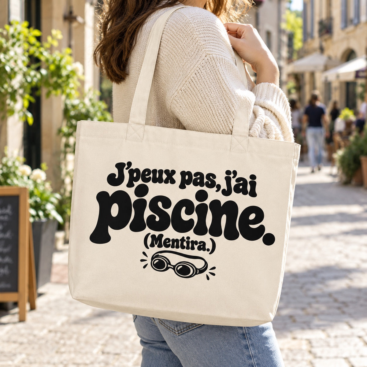Tote bag « J'peux pas, j'ai piscine. (Mentira.) » Premium porté à l'épaule dans une rue piétonne ensoleillée