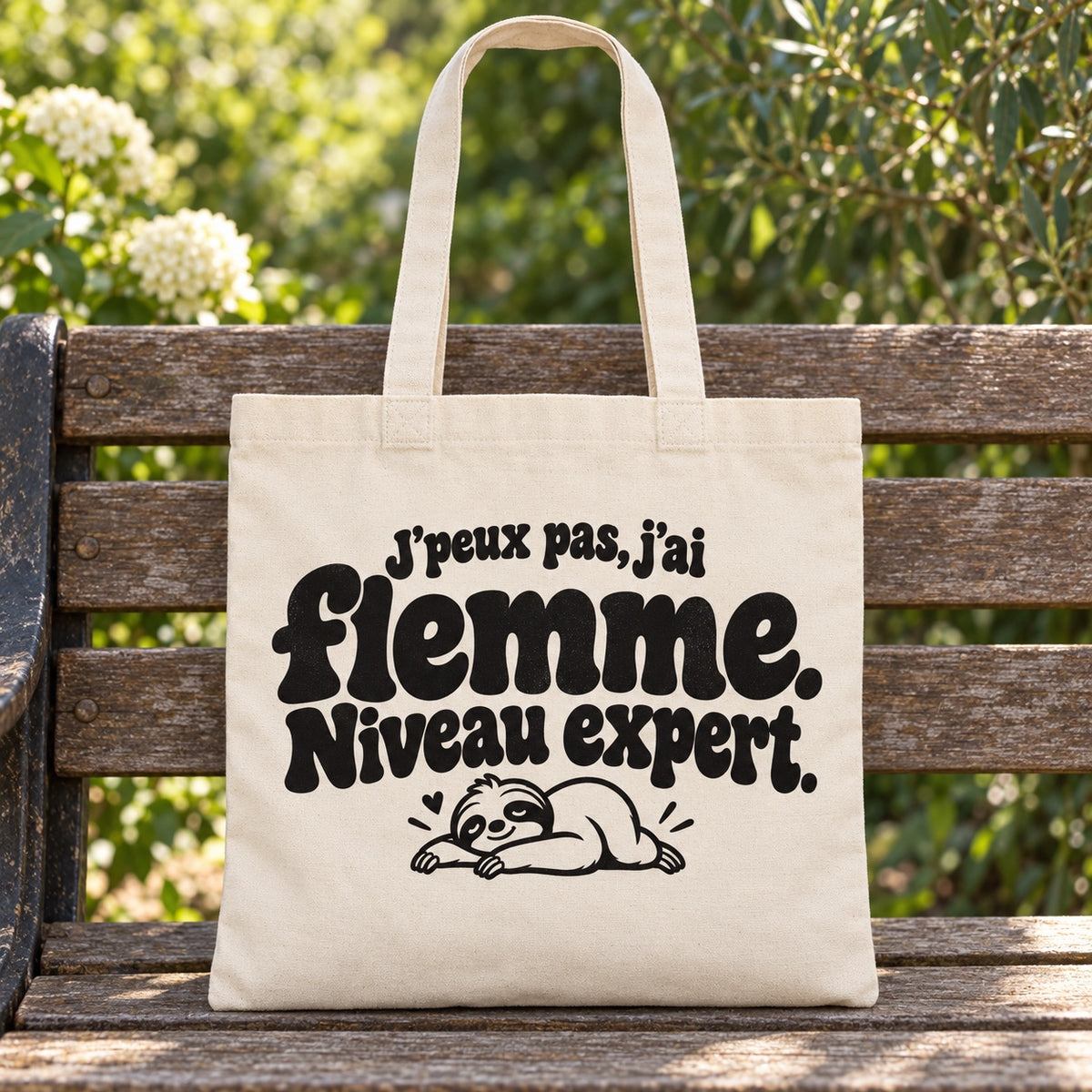 Tote bag « J'peux pas, j'ai flemme. Niveau expert. » Essentiel posé sur un banc en bois dans un jardin