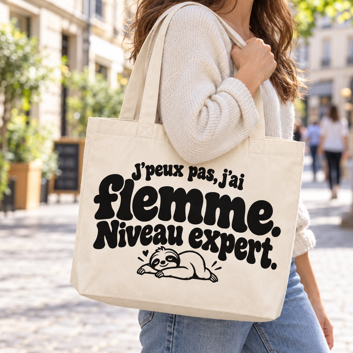 Tote bag « J'peux pas, j'ai flemme. Niveau expert. » Premium porté à l'épaule dans une rue piétonne ensoleillée