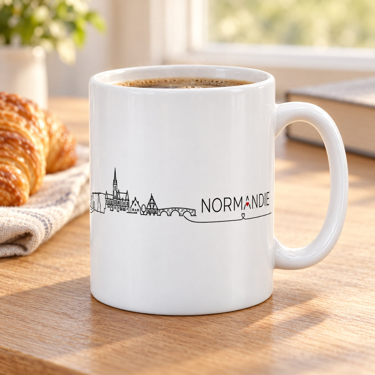 Mug Normandie Skyline « Étretat & Colombages » posé sur une table en bois avec un croissant, pause café