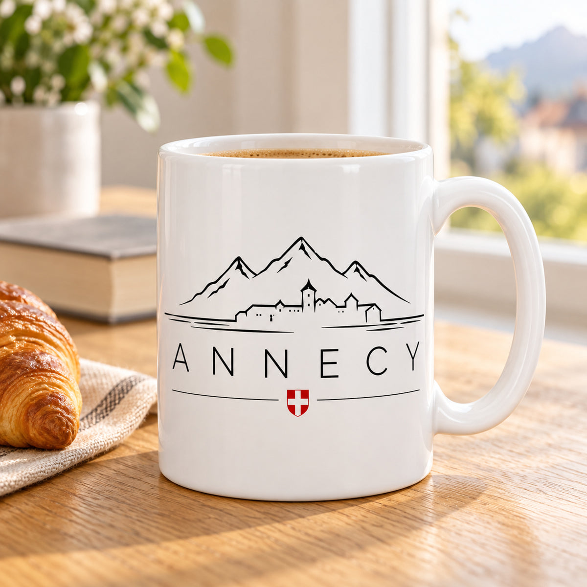 Mug Annecy Skyline « Château & Montagnes » posé sur une table en bois avec un croissant, pause café