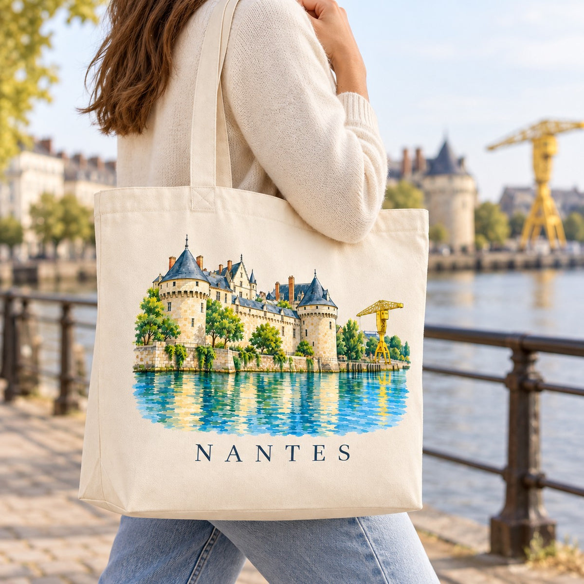 Tote bag Nantes Premium porté à l'épaule le long d'un quai, château et grue jaune en arrière-plan