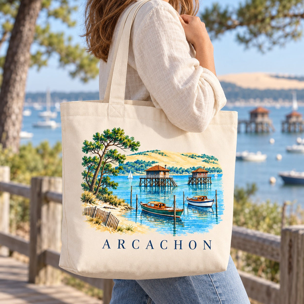 Tote bag Arcachon Premium porté à l'épaule sur une promenade face au Bassin, dune du Pilat au loin