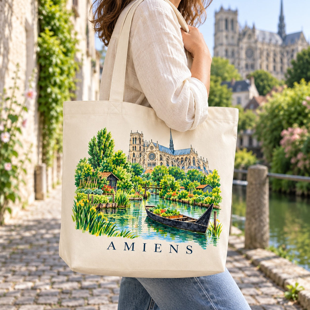 Tote bag Amiens Premium porté à l'épaule dans une ruelle au bord de l'eau, cathédrale en arrière-plan