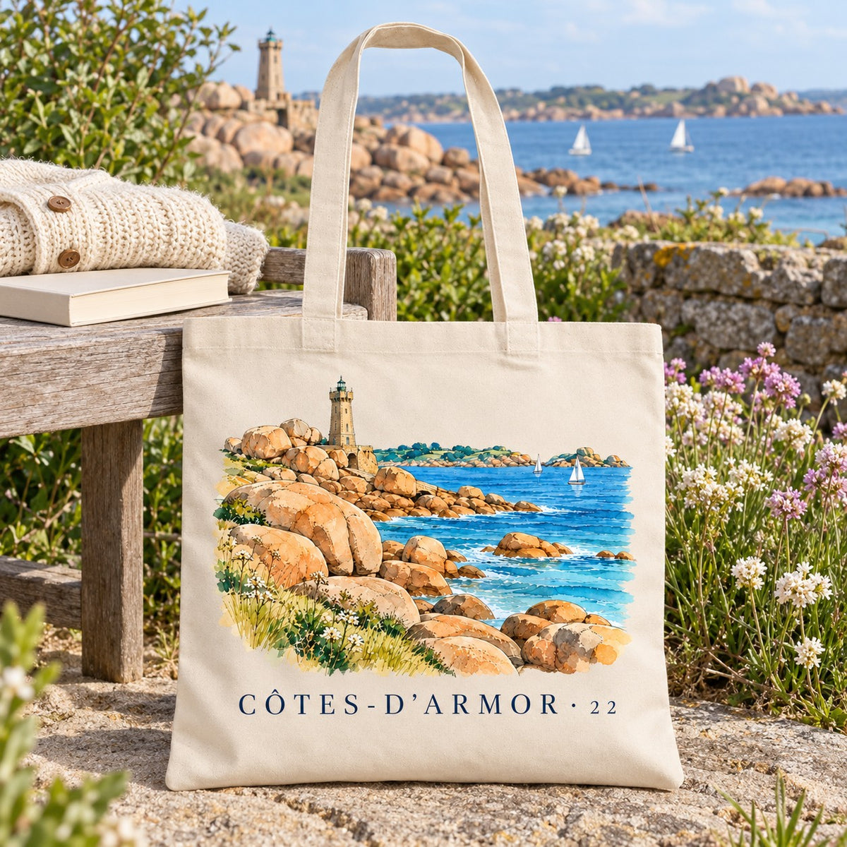 Tote bag Côtes-d'Armor (22) Essentiel posé sur un banc en bois face à la côte, avec un livre et un pull