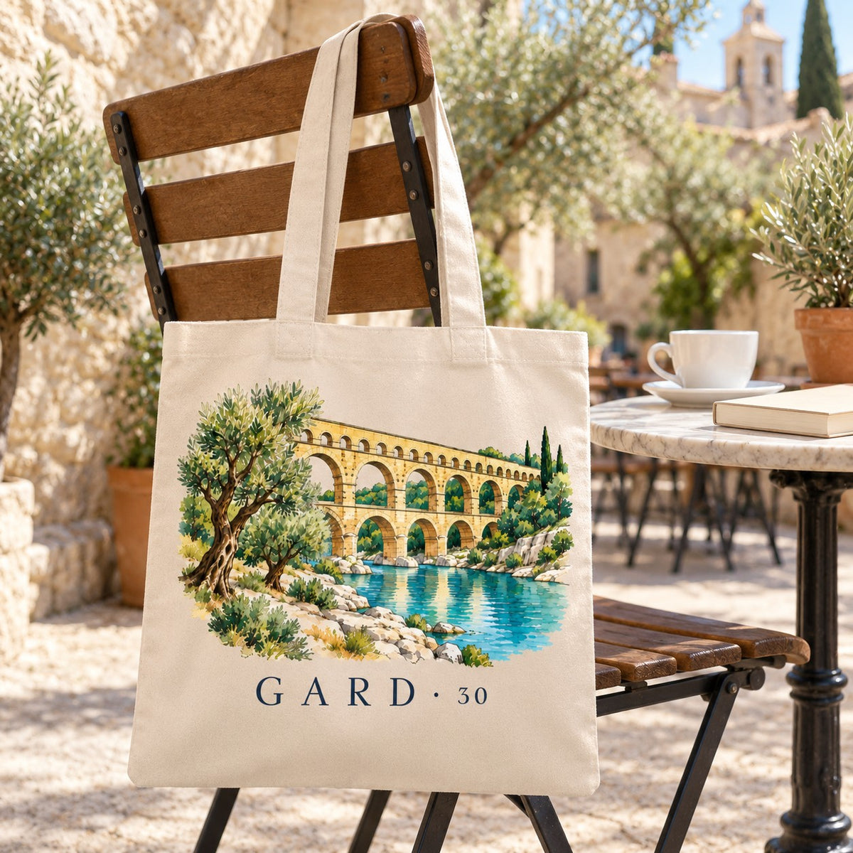 Tote bag Gard (30) Essentiel suspendu à une chaise en terrasse d'un village de pierre, avec un café