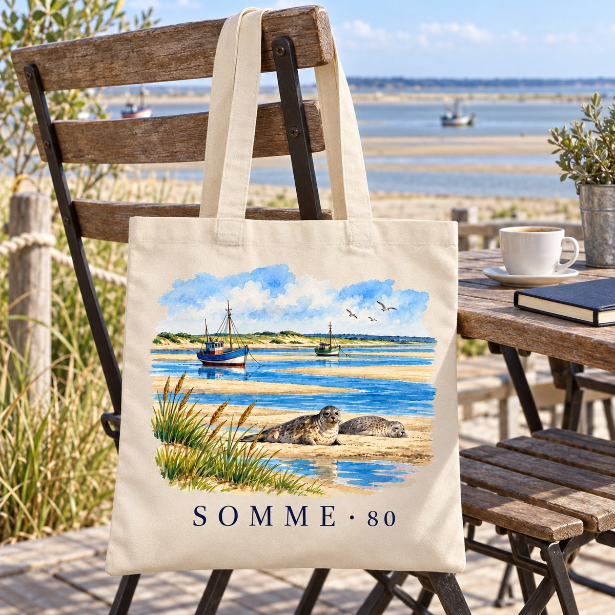 Tote bag Somme (80) Essentiel suspendu à une chaise en terrasse face à la baie, avec un café et un carnet