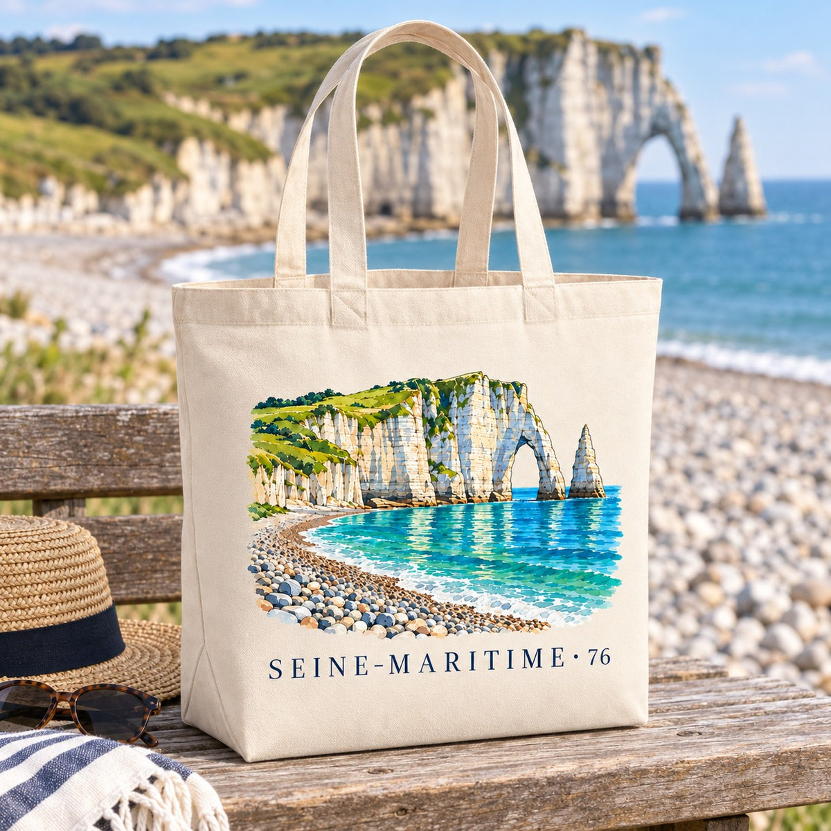 Tote bag Seine-Maritime (76) Premium posé sur un banc de bois face à la plage de galets, avec un chapeau de paille