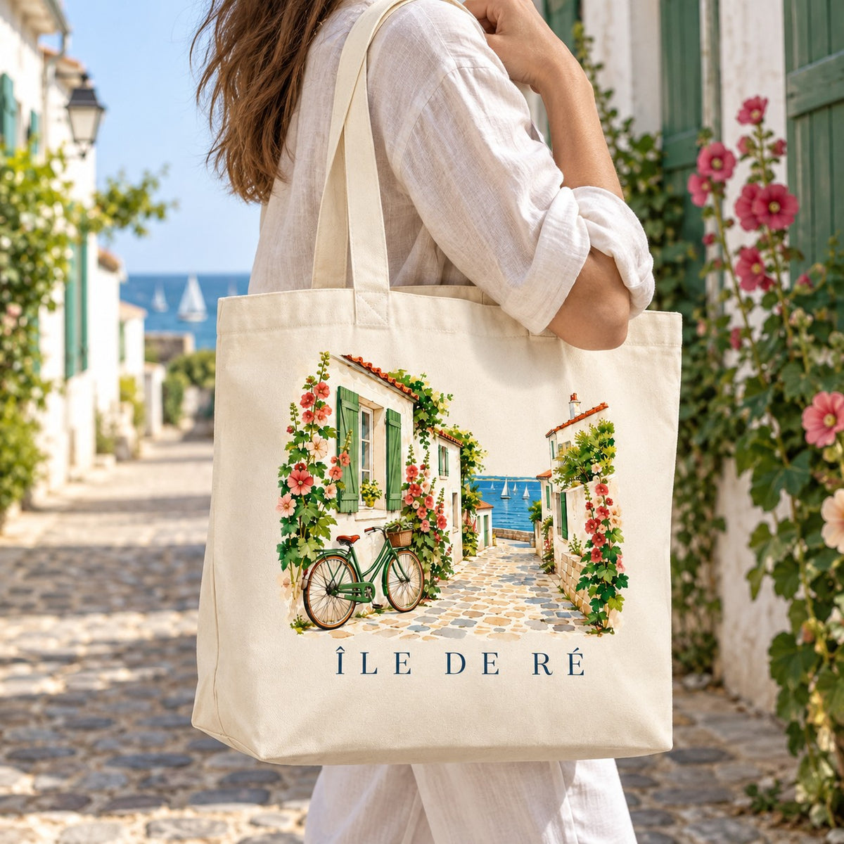 Tote bag Île de Ré Premium porté à l'épaule dans une ruelle bordée de roses trémières