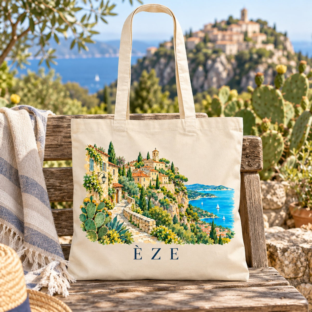 Tote bag Èze Essentiel posé sur un banc de bois entre les cactus, face au village perché