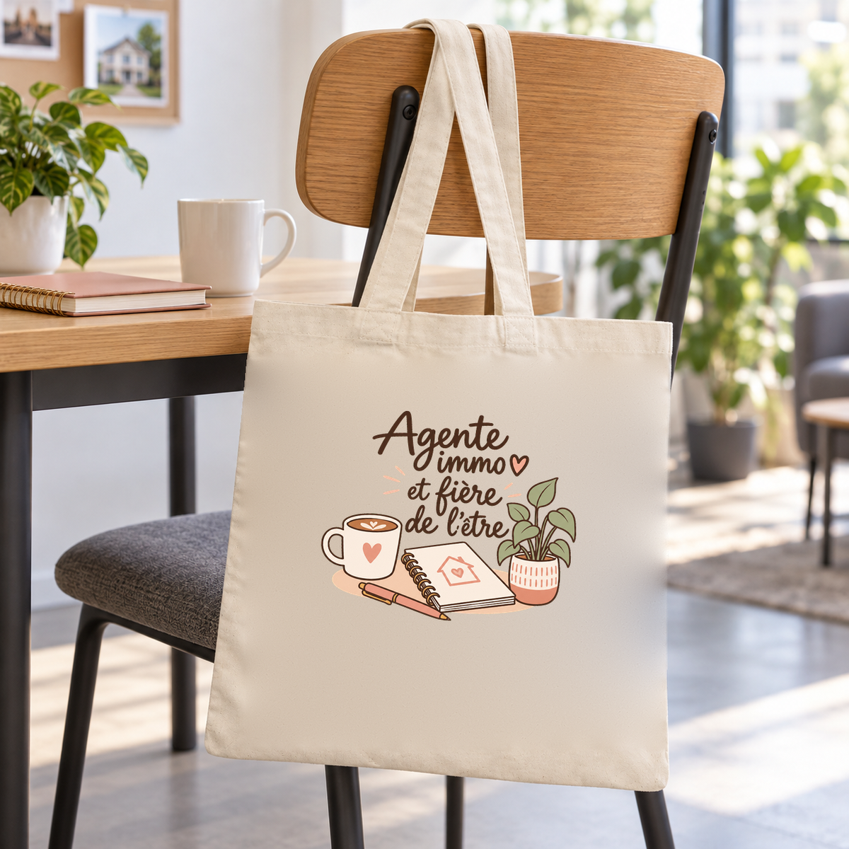 Tote bag agente immobilière « Agente immo et fière de l'être » Essentiel suspendu à une chaise de bureau, à côté d'un mug