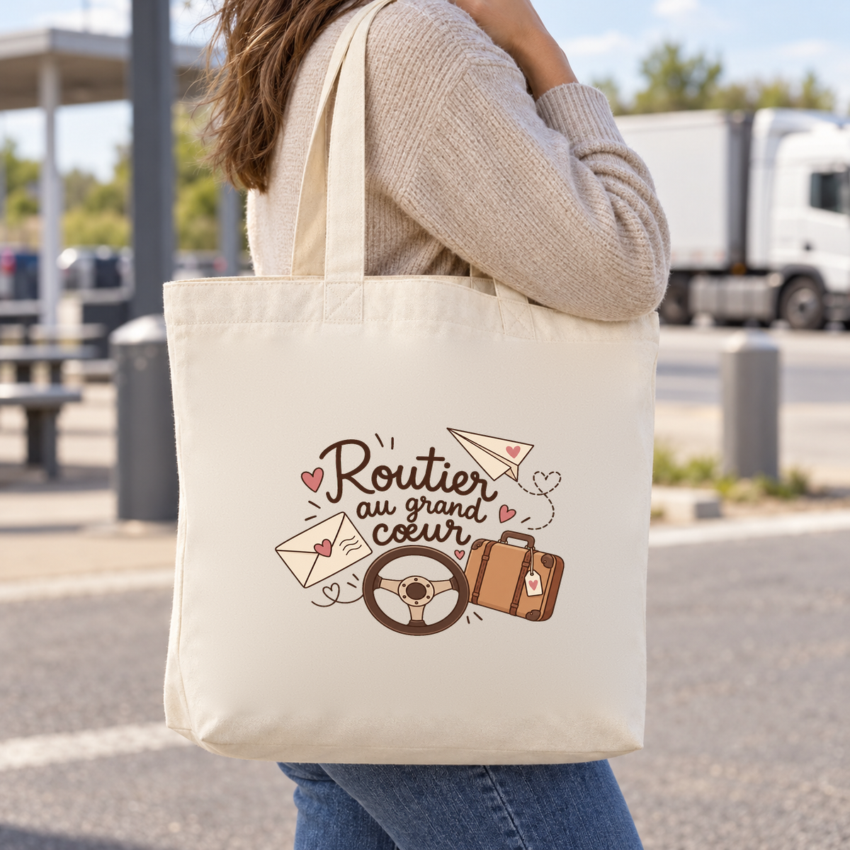 Tote bag chauffeur routier « Routier au grand cœur » Premium porté à l'épaule devant une aire de stationnement de camions