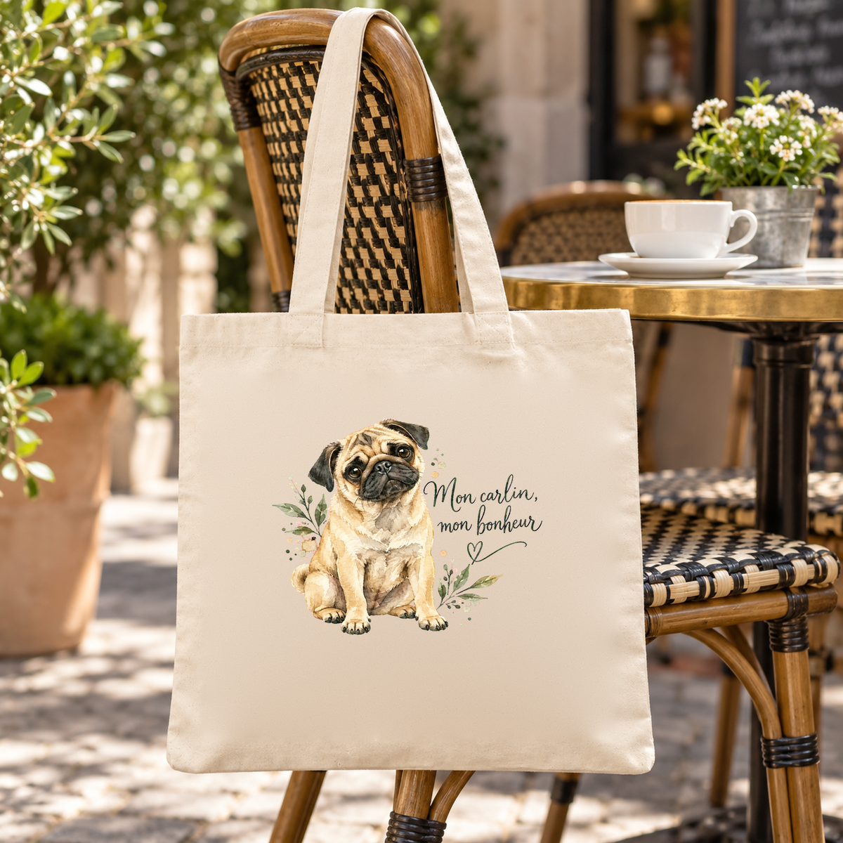 Tote bag carlin « Mon carlin, mon bonheur » Essentiel suspendu à une chaise de terrasse de café, à côté d'une tasse