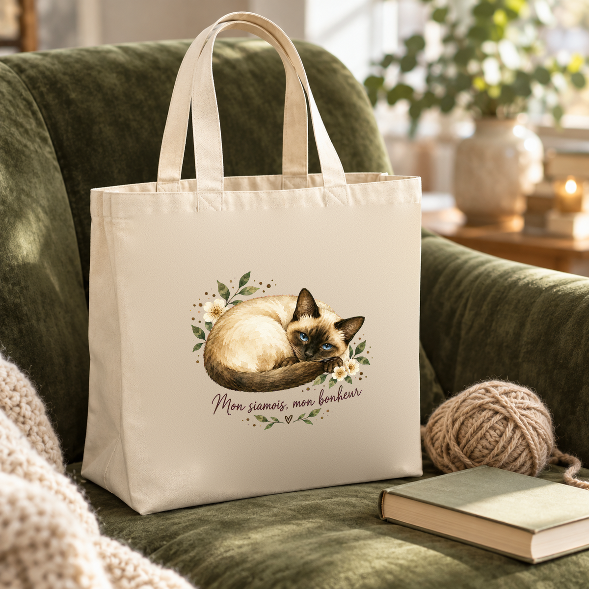 Tote bag siamois « Mon siamois, mon bonheur » Premium posé sur un fauteuil en velours vert, à côté d'une pelote de laine et d'un livre