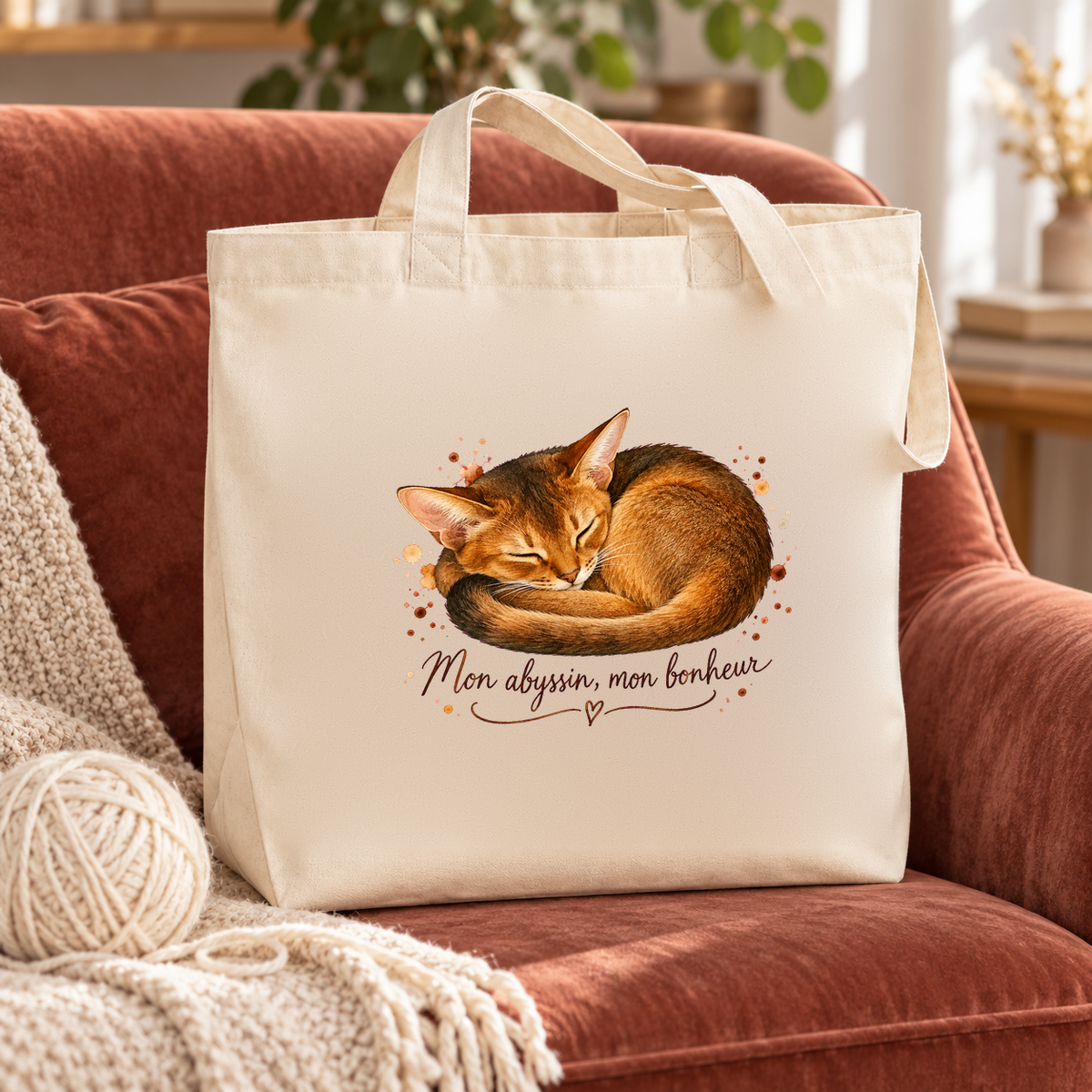 Tote bag abyssin « Mon abyssin, mon bonheur » Premium posé sur un fauteuil en velours rose, à côté d'un plaid et d'une pelote de laine