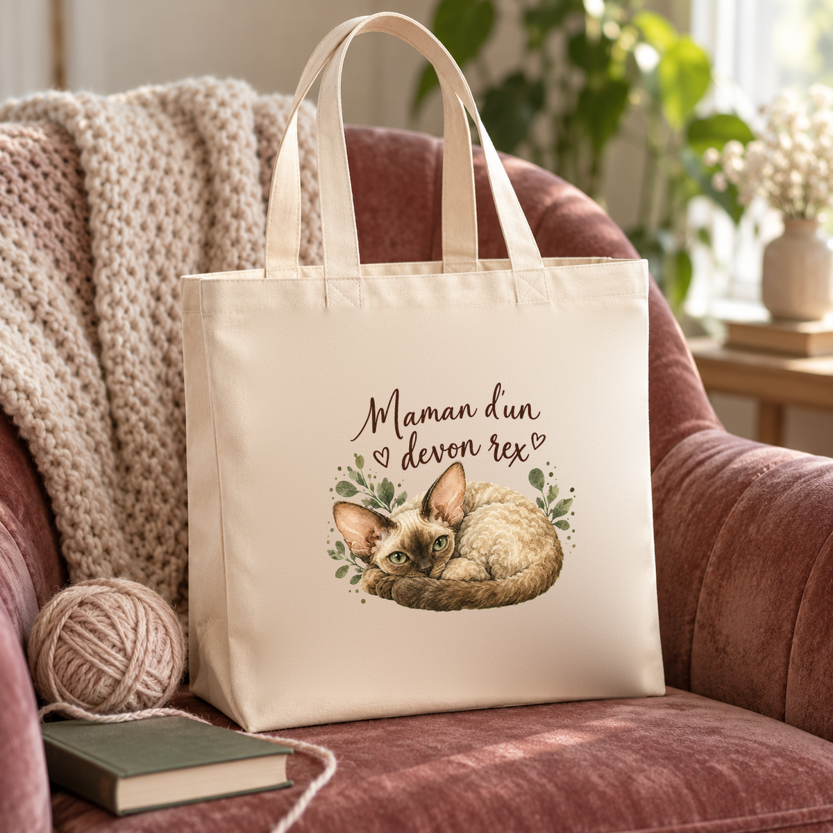 Tote bag devon rex « Maman d'un devon rex » Premium posé sur un fauteuil en velours rose, à côté d'une pelote de laine et d'un livre