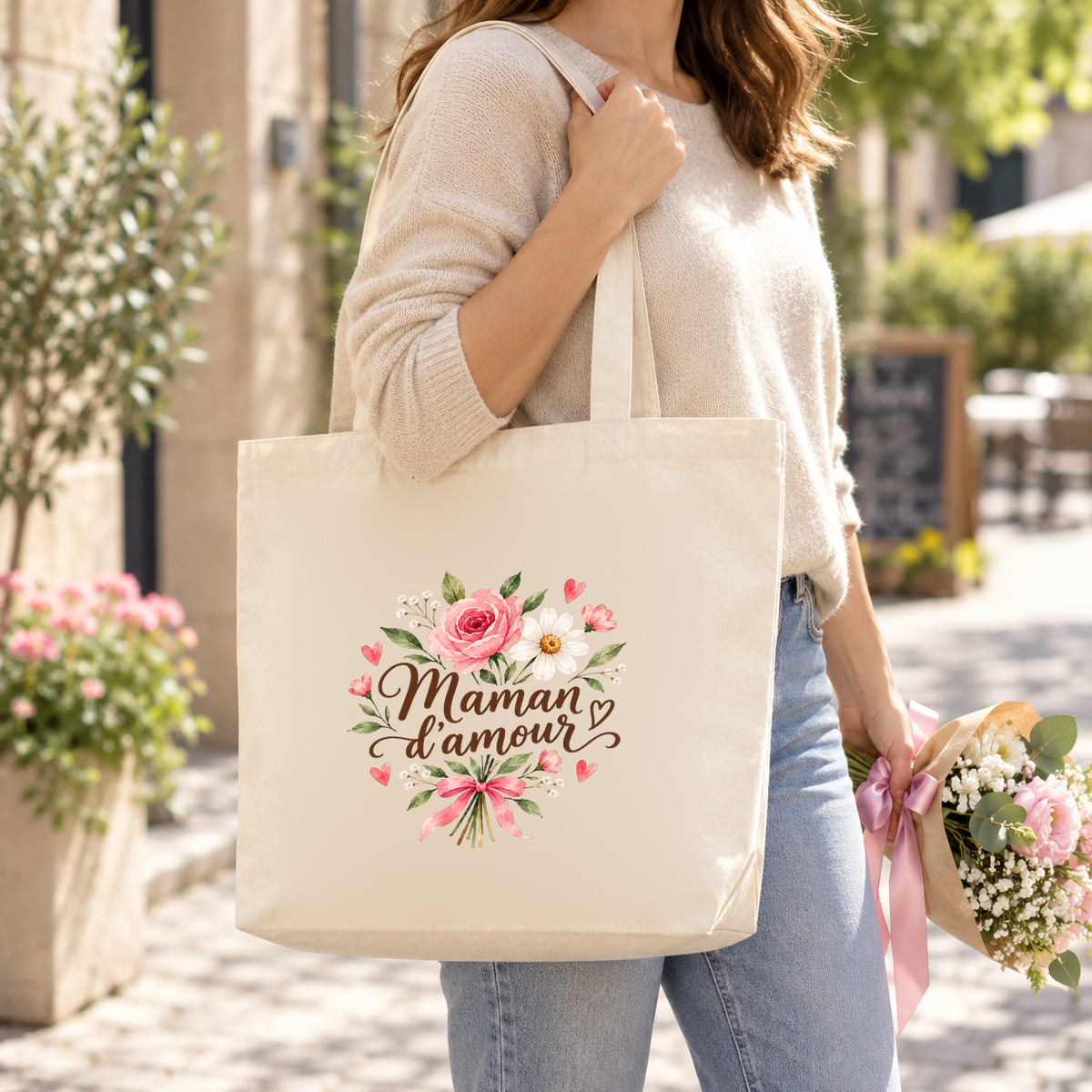 Tote bag maman « Maman d'amour » Premium porté à l'épaule dans une rue fleurie, un bouquet de fleurs à la main