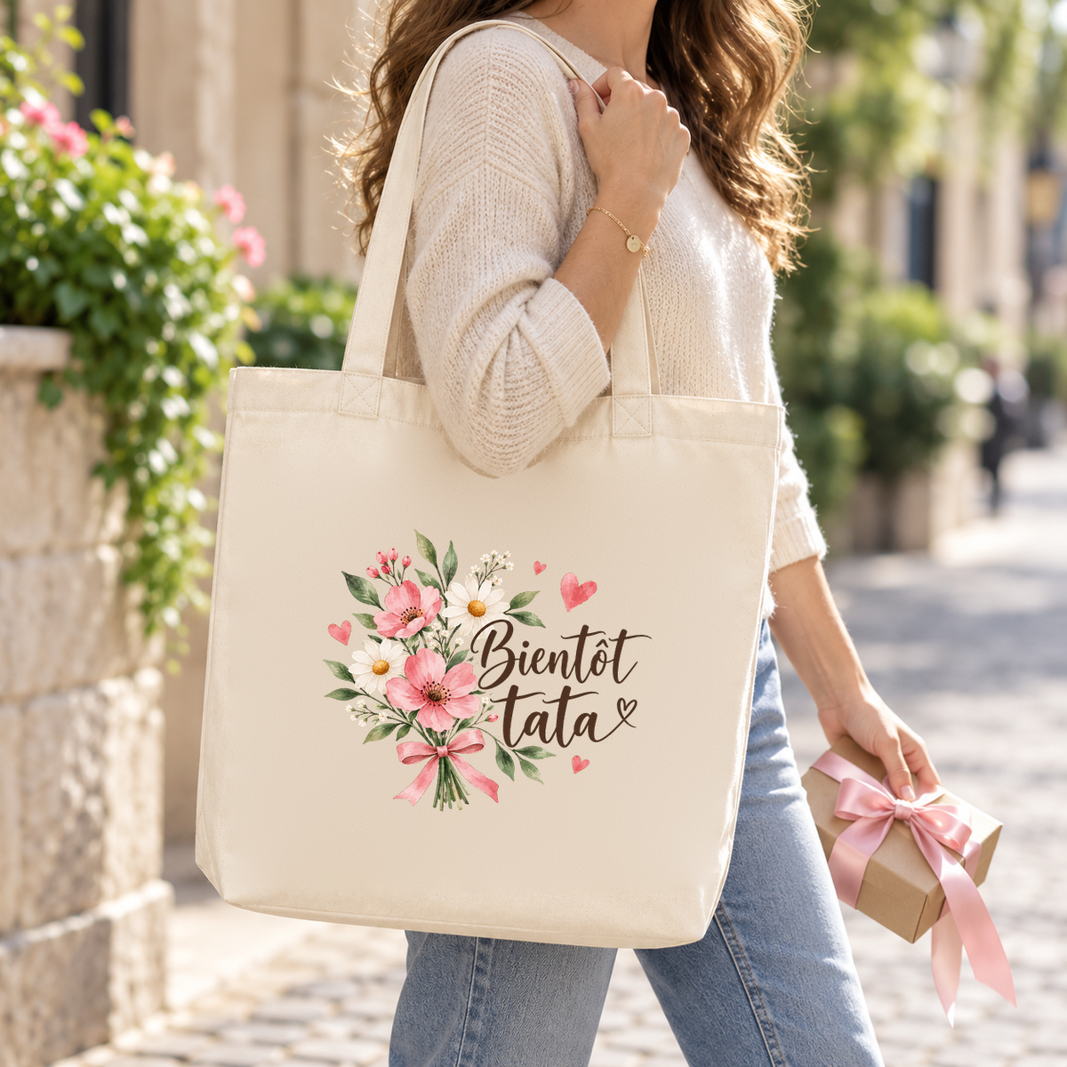 Tote bag tata « Bientôt tata » Premium porté à l'épaule dans une rue ensoleillée, un cadeau à la main