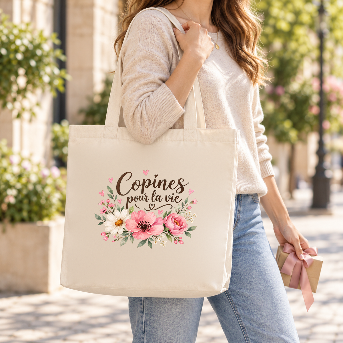 Tote bag amitié « Copines pour la vie » Premium porté à l'épaule dans une rue fleurie, un bouquet de fleurs à la main