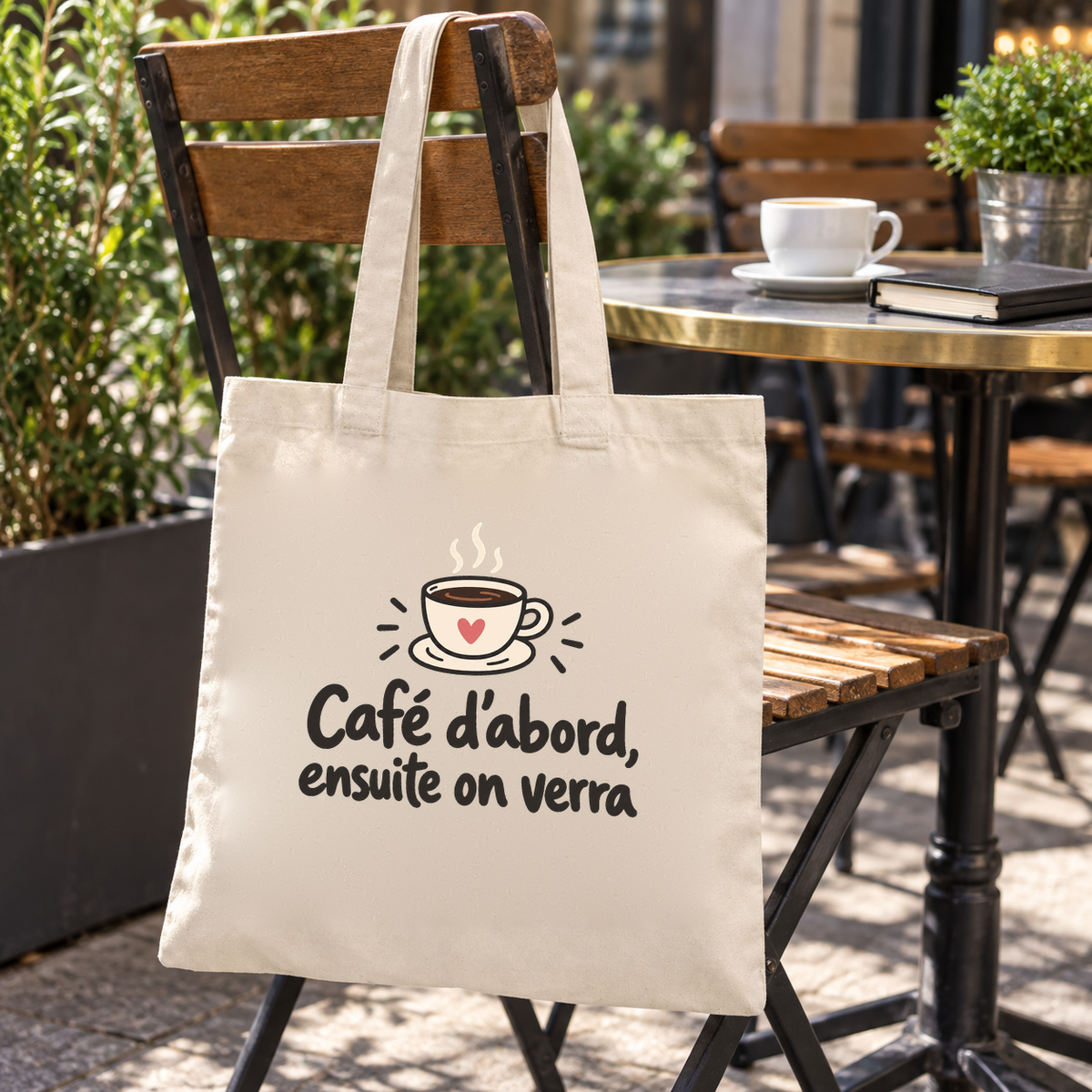Tote bag « Café d'abord, ensuite on verra » Essentiel suspendu à une chaise de bistrot en terrasse