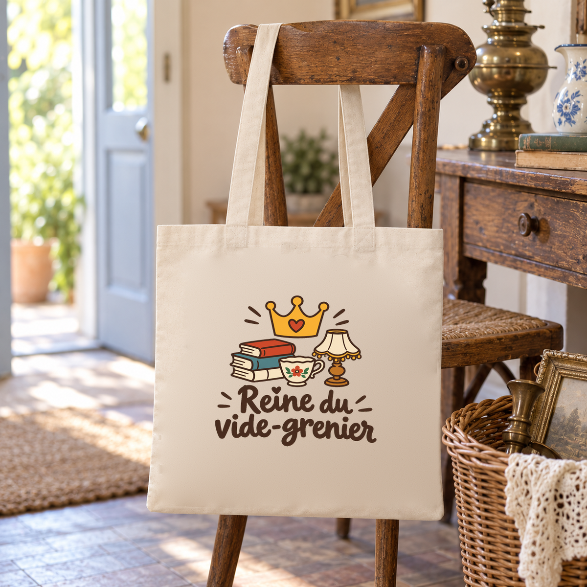 Tote bag « Reine du vide-grenier » Essentiel suspendu à une chaise près d'une porte, à côté d'un panier en osier