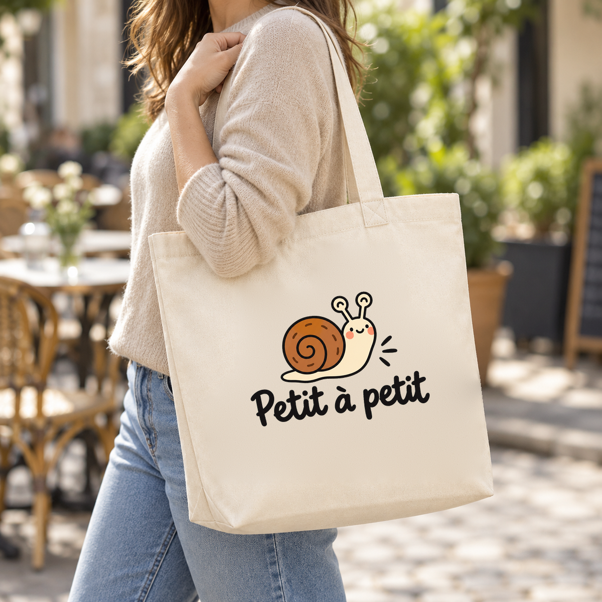 Tote bag « Petit à petit » Premium porté à l'épaule dans une rue ensoleillée, devant une terrasse
