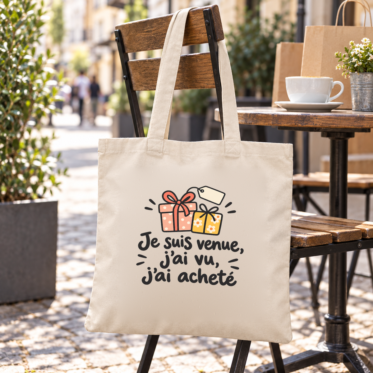 Tote bag « Je suis venue, j'ai vu, j'ai acheté » Essentiel suspendu à une chaise de bistrot en terrasse, à côté de sacs en papier kraft