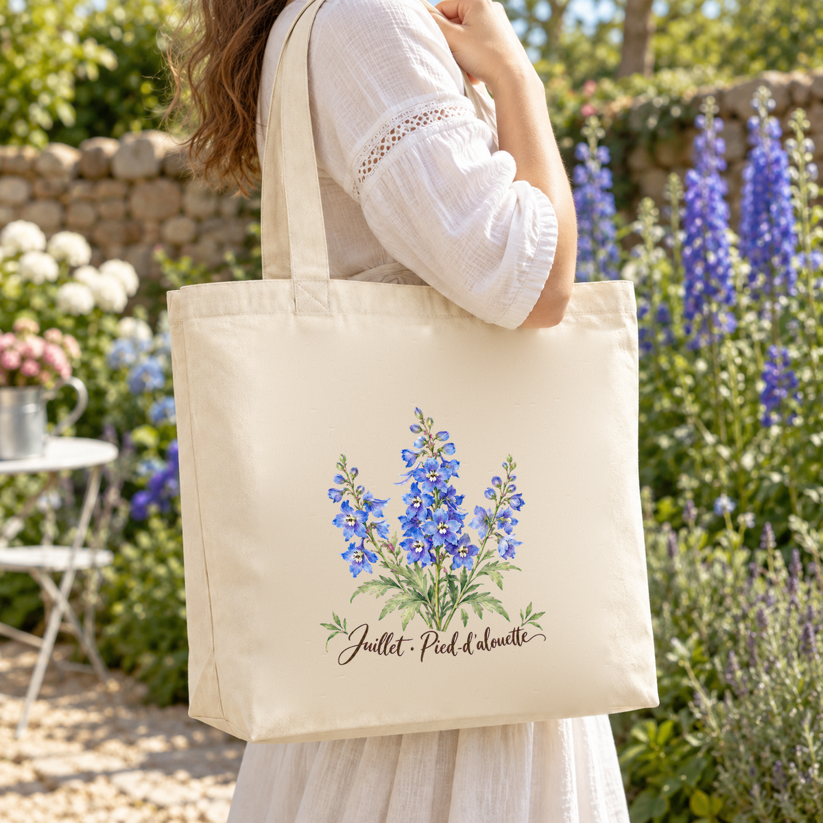Tote bag fleur de naissance « Juillet · Pied-d'alouette » Premium porté à l'épaule dans un jardin fleuri de pieds-d'alouette