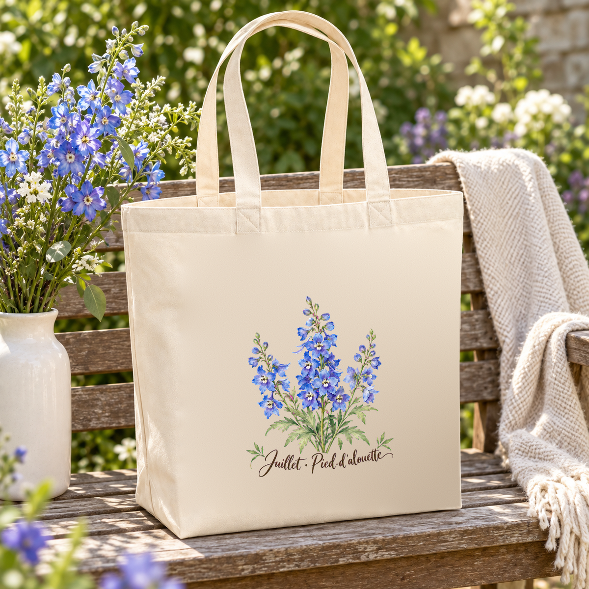 Tote bag fleur de naissance « Juillet · Pied-d'alouette » Premium posé sur un banc de jardin, à côté d'un vase de pieds-d'alouette et d'un plaid
