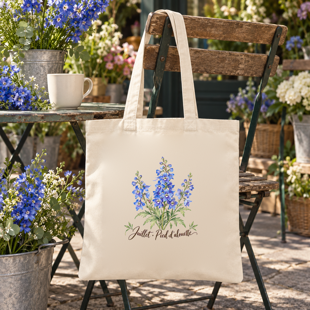 Tote bag fleur de naissance « Juillet · Pied-d'alouette » Essentiel suspendu à une chaise de bistrot en terrasse, à côté d'une tasse et de pieds-d'alouette