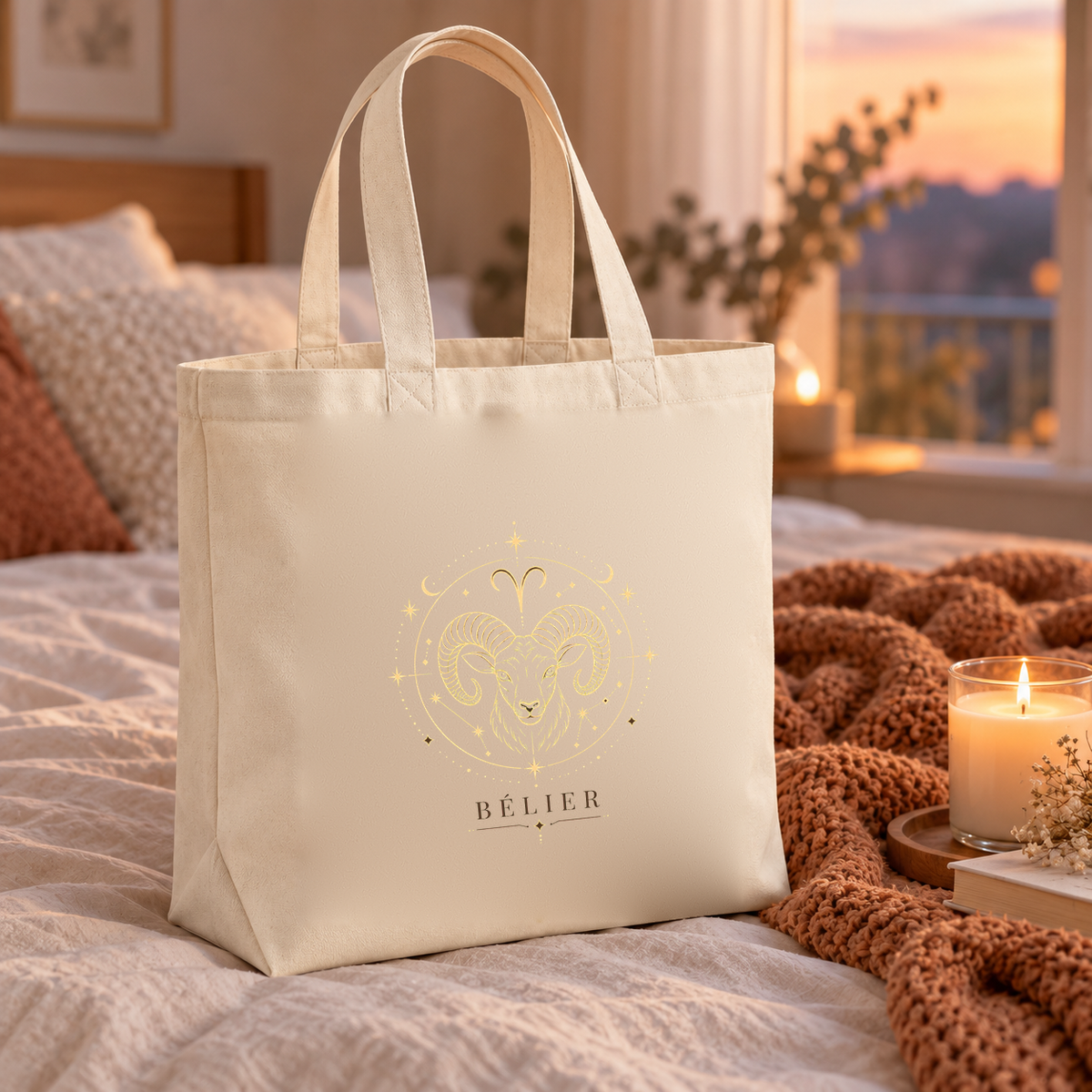Tote bag signe astrologique « BÉLIER » Premium posé sur un lit, à côté d'un plaid et d'une bougie