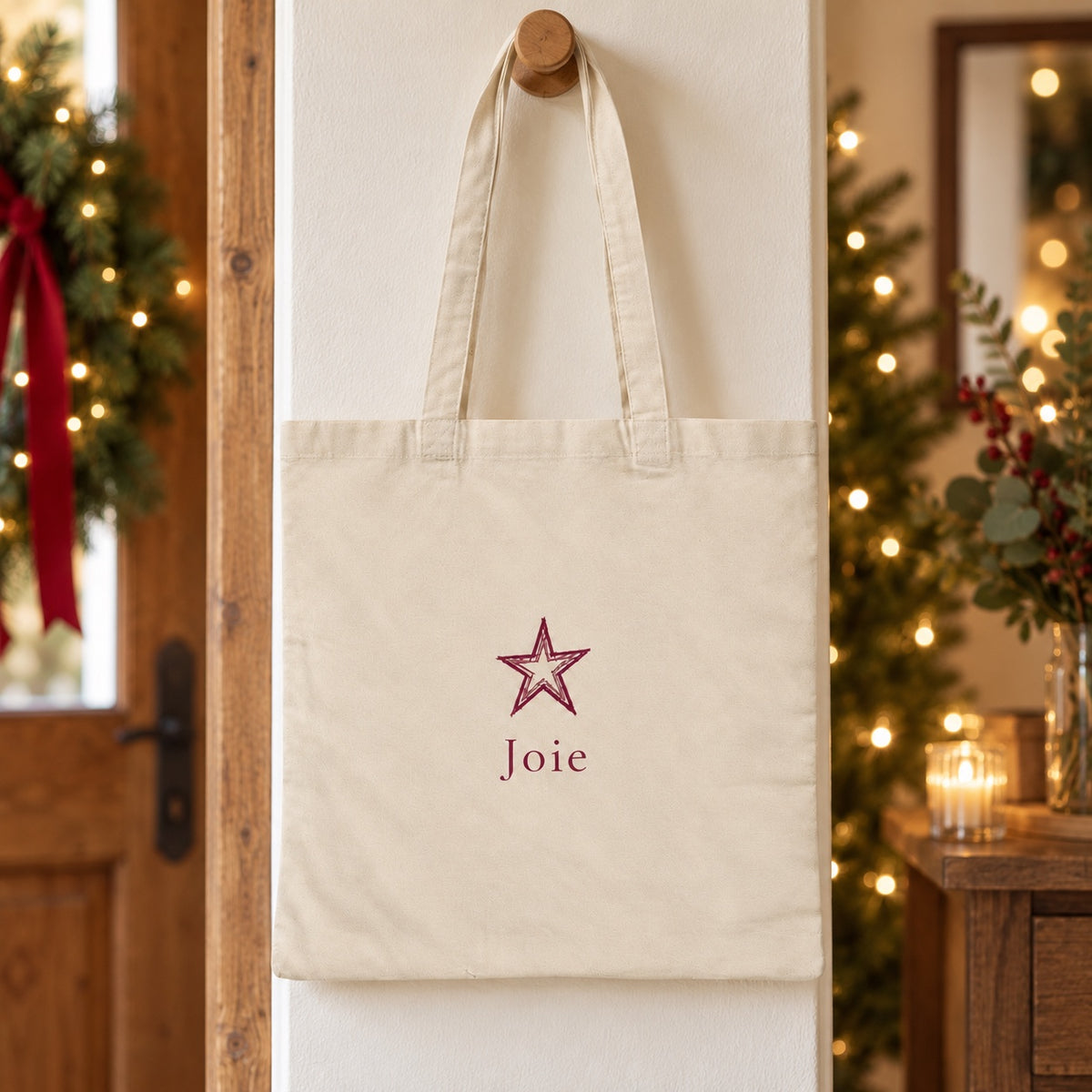 Tote bag de Noël « Joie » Essentiel suspendu à une patère dans une entrée décorée pour Noël