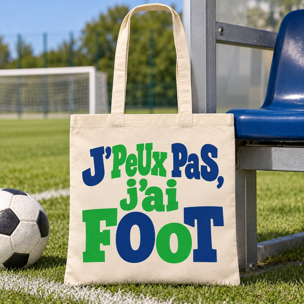 Tote bag « J'peux pas, j'ai foot » Essentiel posé sur la pelouse près d'un ballon et d'un siège de tribune