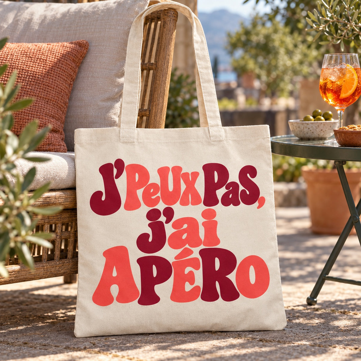 Tote bag « J'peux pas, j'ai apéro » Essentiel appuyé contre un fauteuil de terrasse à l'heure de l'apéritif