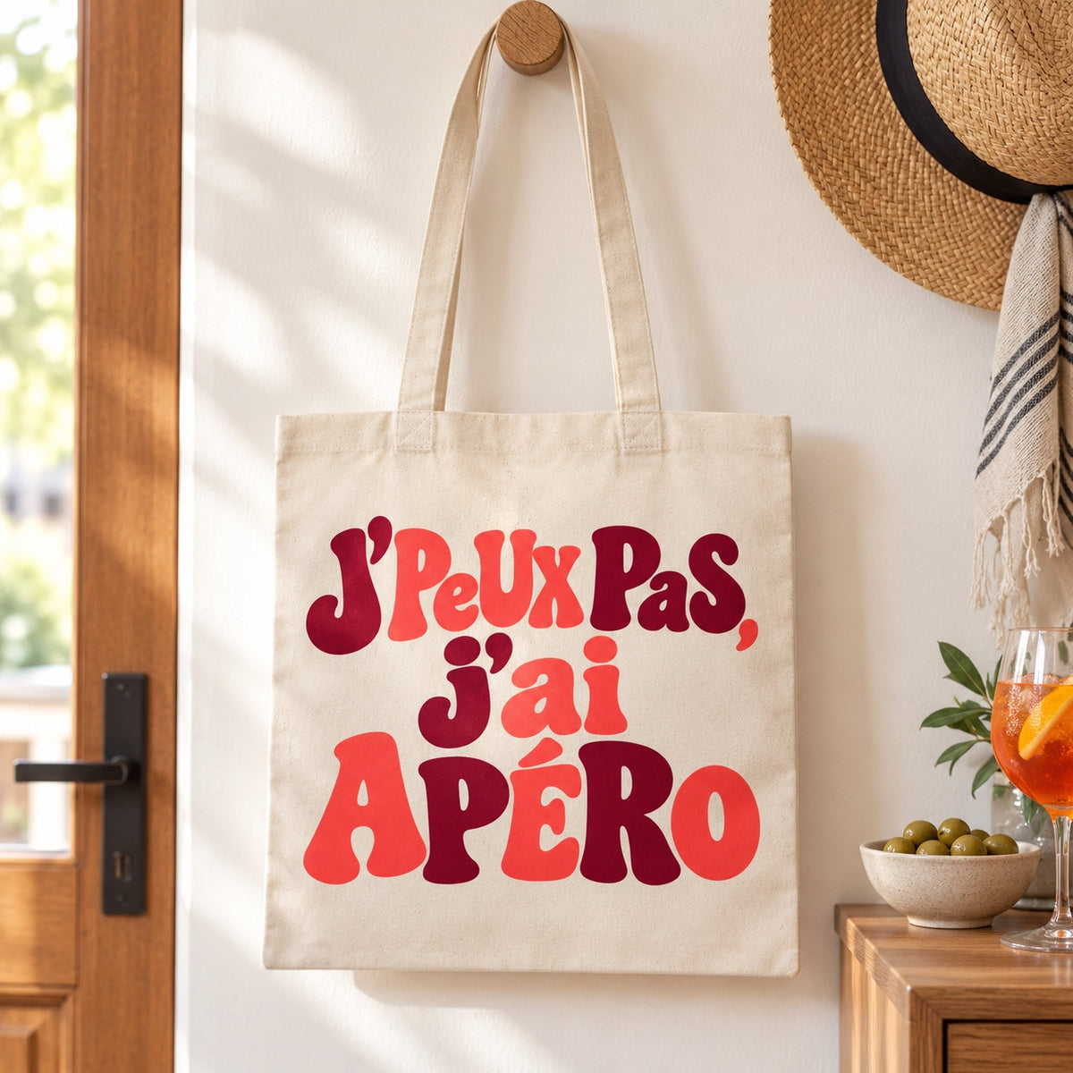 Tote bag « J'peux pas, j'ai apéro » Essentiel suspendu à une patère dans une entrée, à côté d'un chapeau de paille