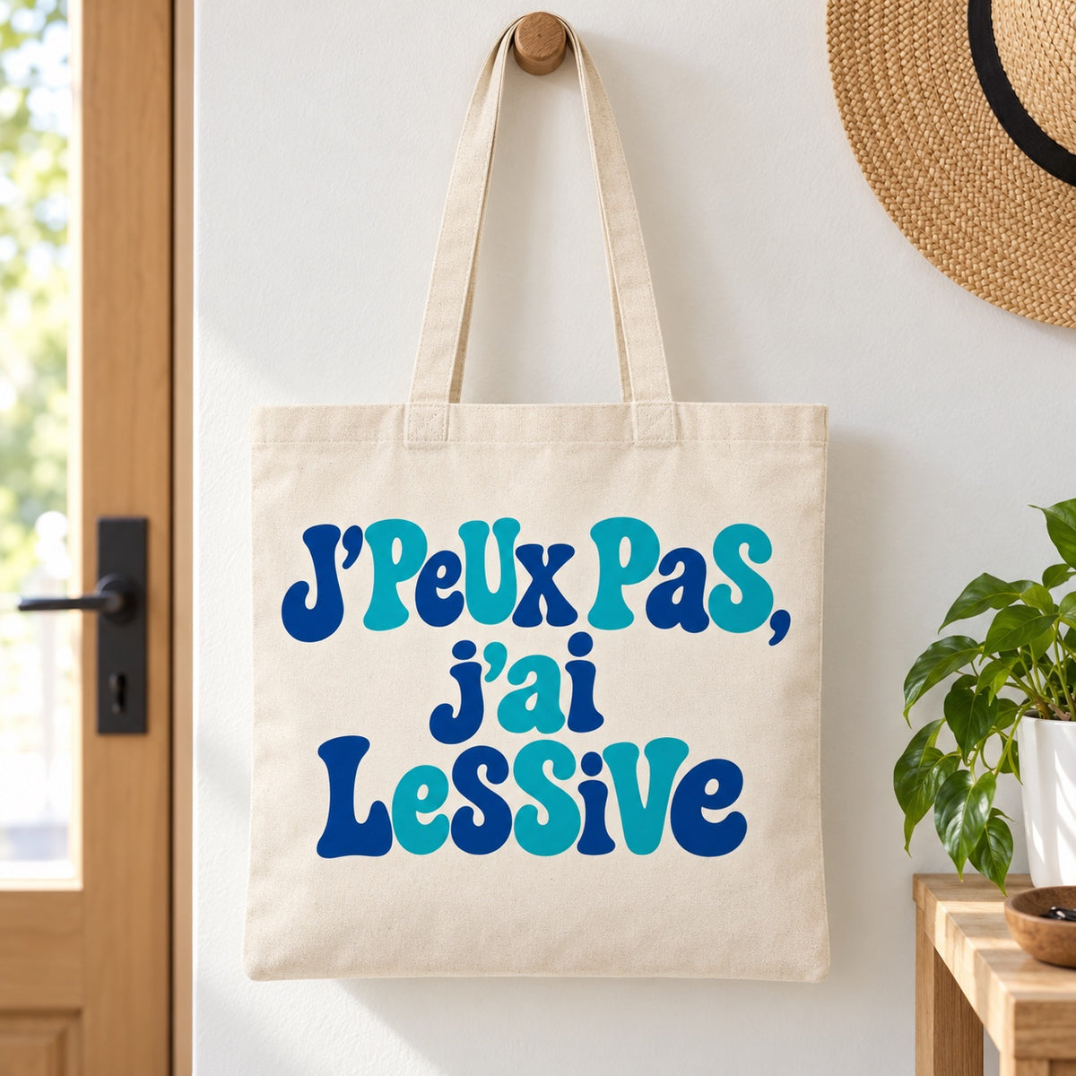 Tote bag « J'peux pas, j'ai lessive » Essentiel suspendu à une patère dans une entrée