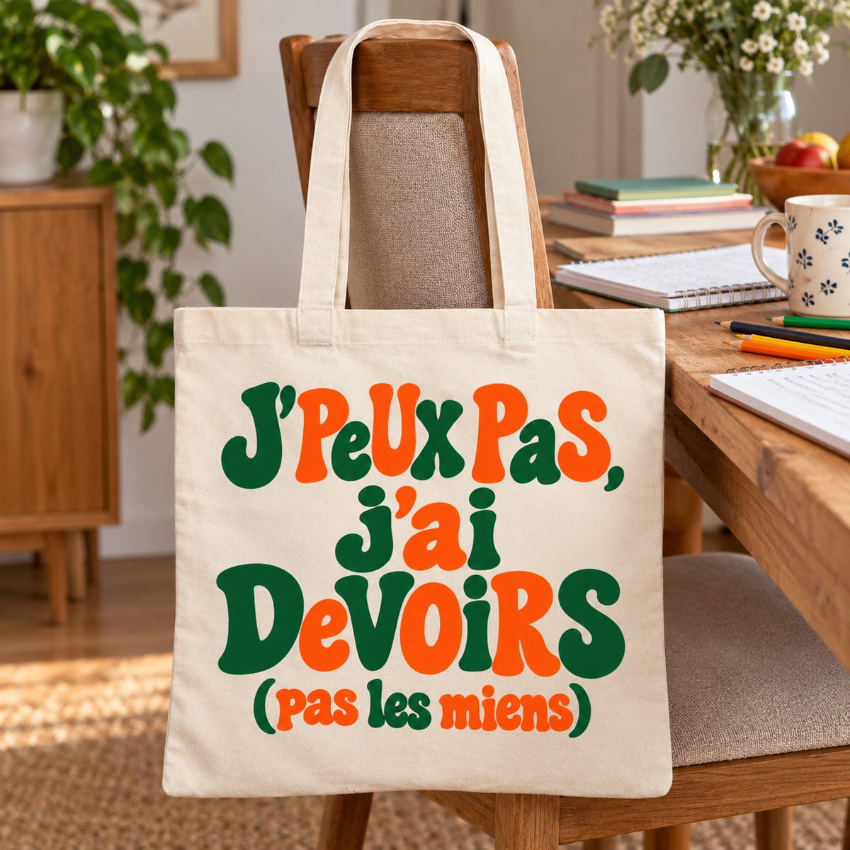 Tote bag « J'peux pas, j'ai devoirs (pas les miens) » Essentiel appuyé contre une chaise près d'une table de devoirs