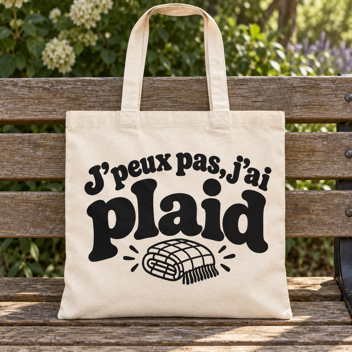 Tote bag « J'peux pas, j'ai plaid » Essentiel posé sur un banc en bois dans un jardin