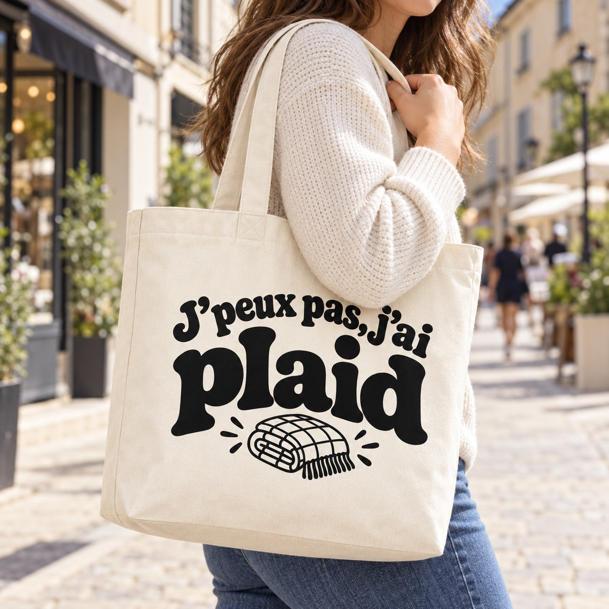 Tote bag « J'peux pas, j'ai plaid » Premium porté à l'épaule dans une rue piétonne ensoleillée