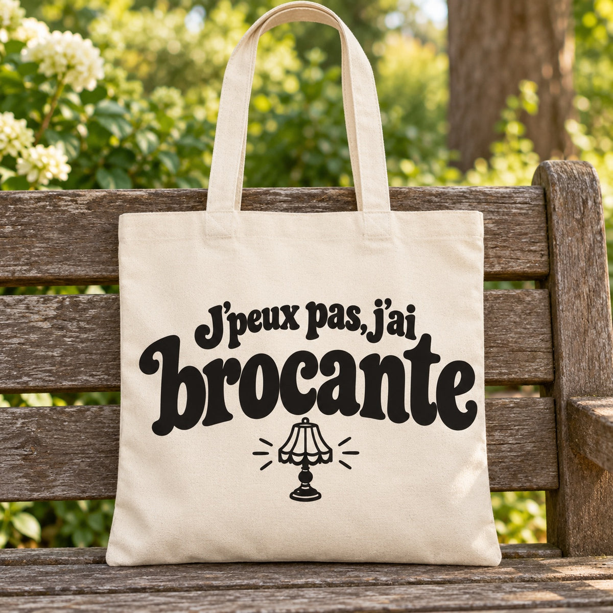 Tote bag « J'peux pas, j'ai brocante » Essentiel posé sur un banc en bois dans un jardin