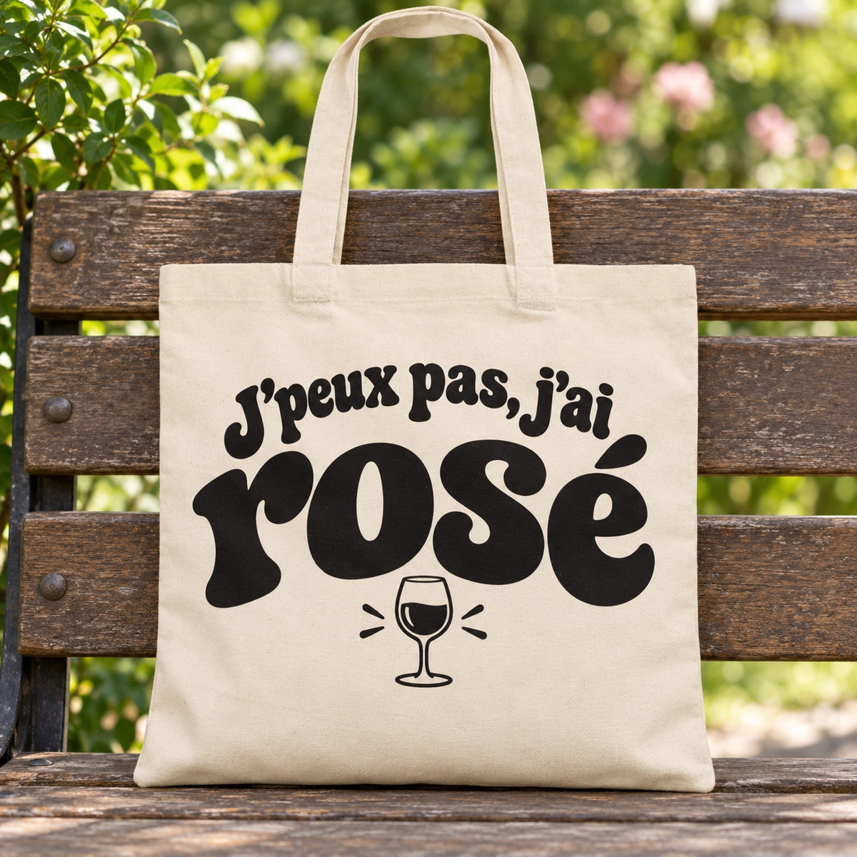 Tote bag « J'peux pas, j'ai rosé » Essentiel posé sur un banc en bois dans un jardin