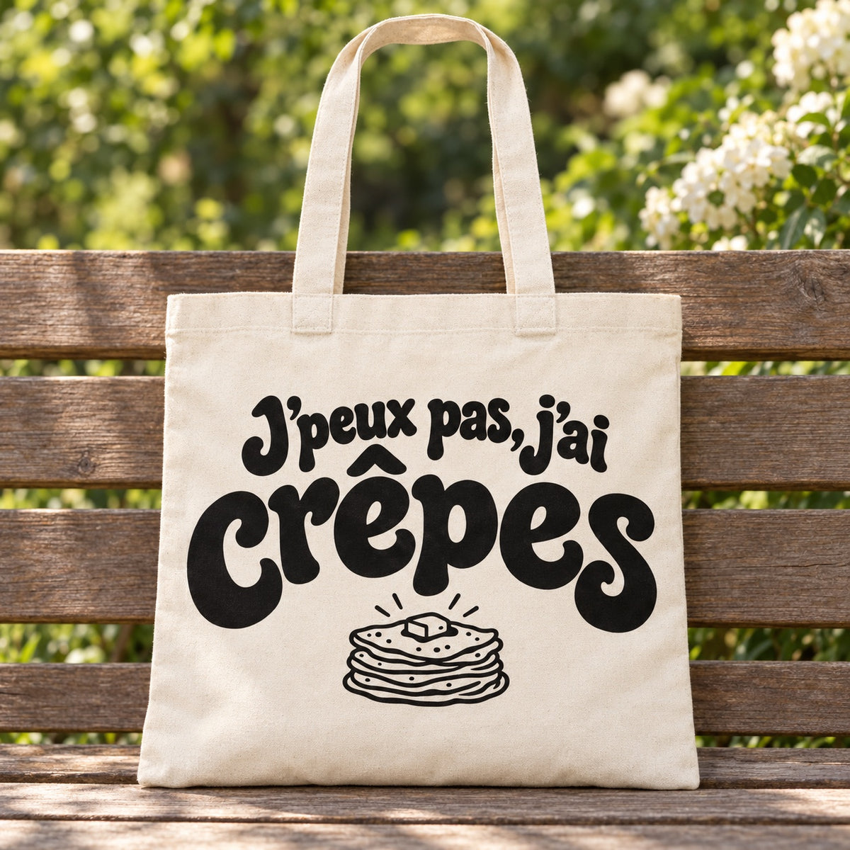 Tote bag « J'peux pas, j'ai crêpes » Essentiel posé sur un banc en bois dans un jardin